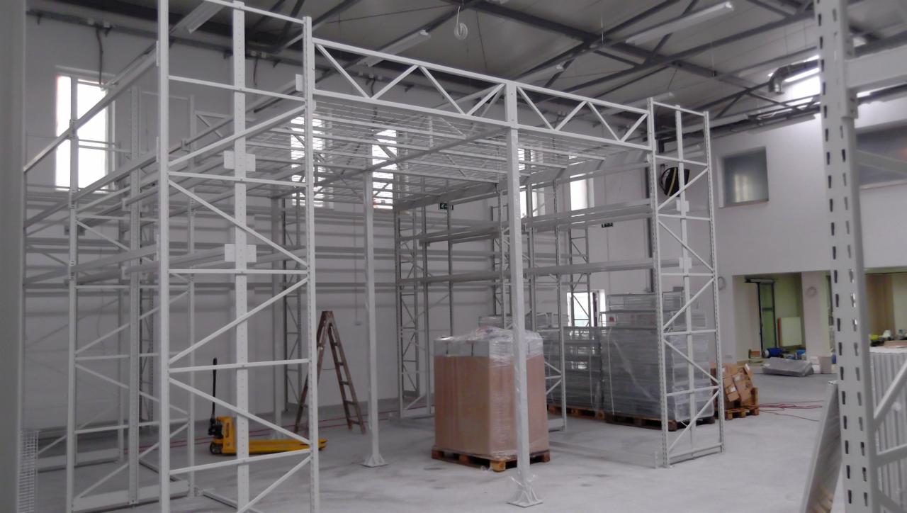 Montaż białych, metalowych regałów magazynowych z widocznymi elementami konstrukcyjnymi, drabina i paleta z towarem w tle, hala magazynowa w trakcie wyposażania.