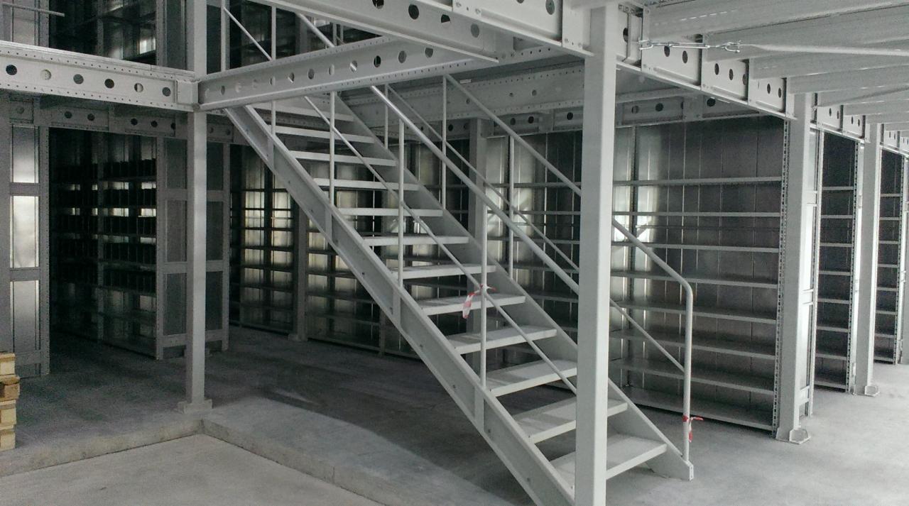 Metalowe schody prowadzące na antresolę z rzędami pustych, metalowych półek w hali magazynowej.