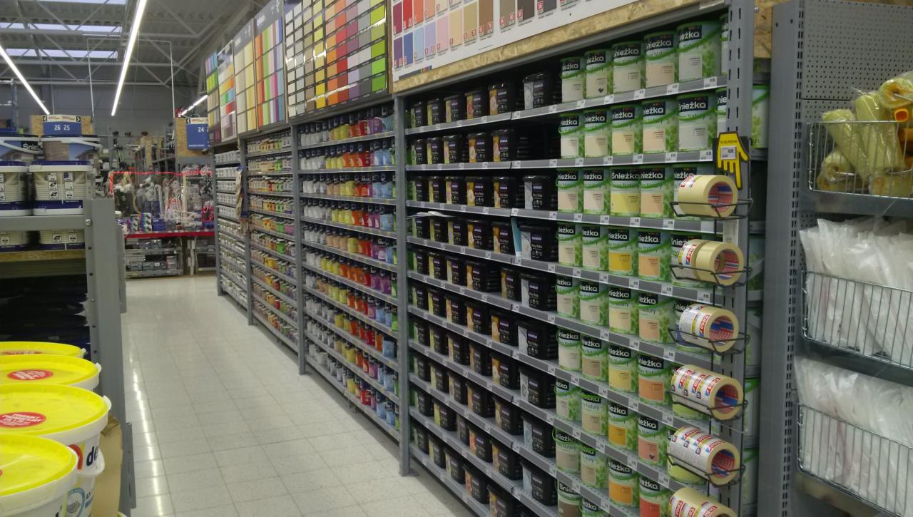 Wnętrze sklepu z szerokim wyborem farb i akcesoriów malarskich na metalowych regałach, widok na alejki z produktami, w tym puszki farb w różnych kolorach i taśmy malarskie.
