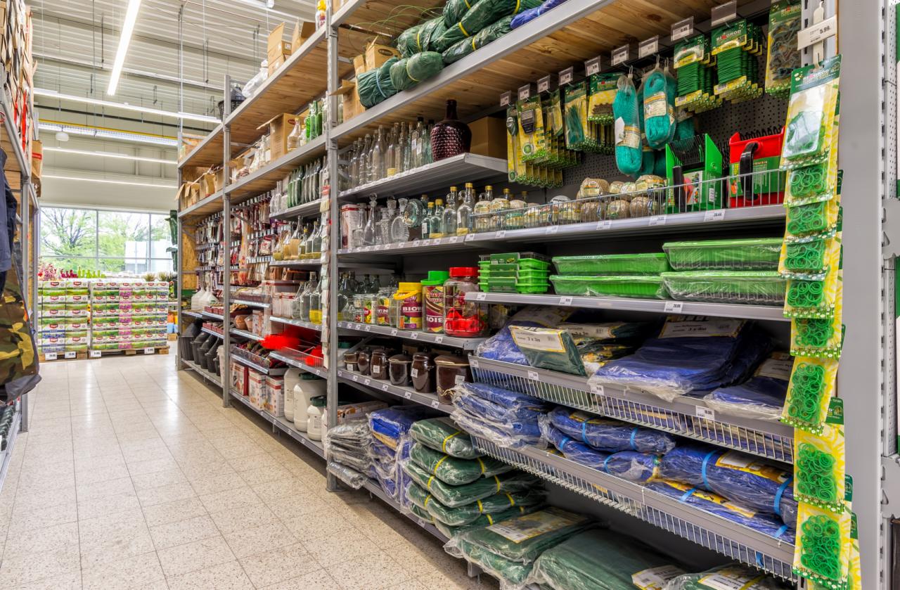 Pełne półki metalowych regałów w sklepie z różnorodnym asortymentem: butelki, słoiki, pojemniki, sznurki, siatki i plandeki w foliowych opakowaniach.