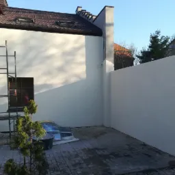 Świeżo otynkowana elewacja budynku i muru ogrodzeniowego w kolorze jasnej szarości, widoczne rusztowanie, małe drzewko iglaste w donicy i kostka brukowa na podwórku.