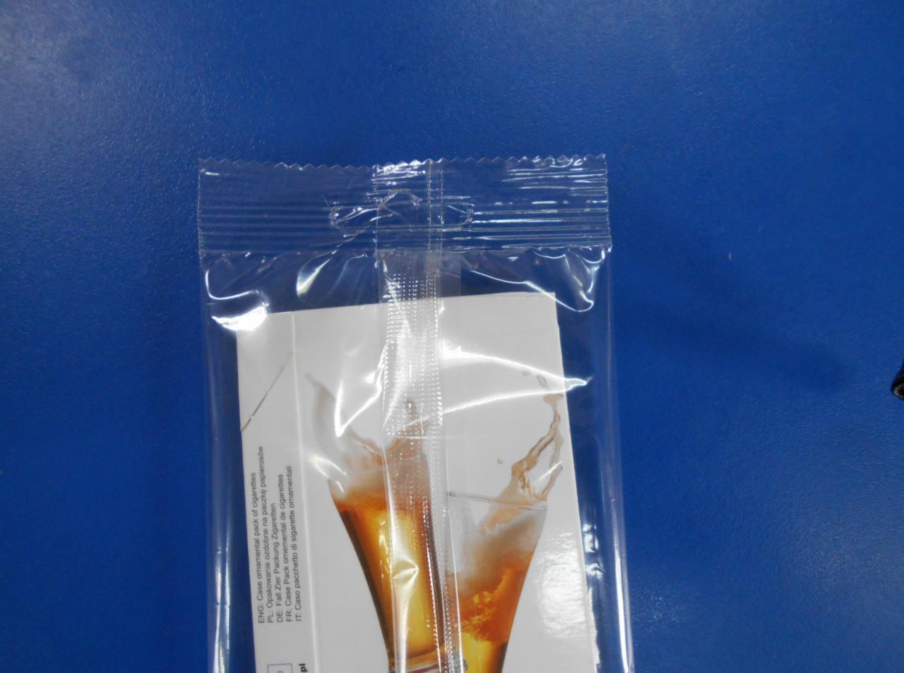 Przezroczyste, zgrzewane opakowanie foliowe zawierające kartonik z nadrukiem przedstawiającym kieliszek z napojem, widoczne tłumaczenia nazwy produktu na różne języki.