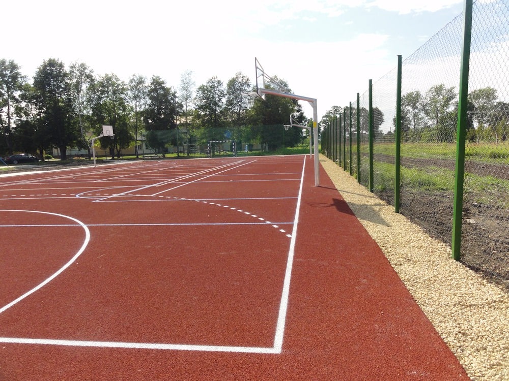 Czerwona nawierzchnia boiska wielofunkcyjnego z białymi liniami, kosz do koszykówki z metalową konstrukcją, ogrodzenie z siatki i zielonych słupków.