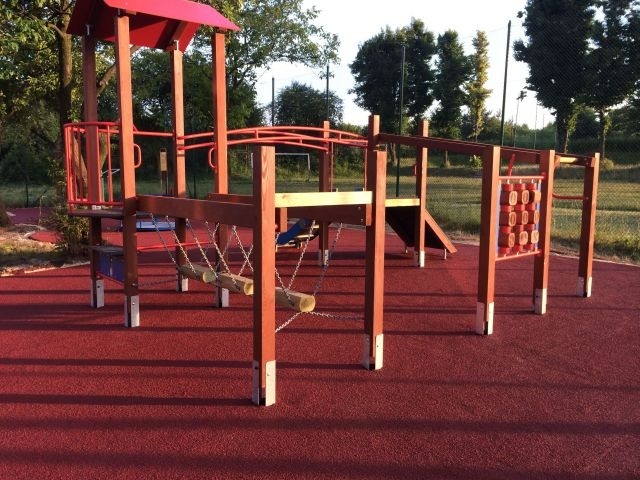 Drewniana konstrukcja zabawowa z wieżą, mostkiem linowym i ścianką wspinaczkową na czerwonej, bezpiecznej nawierzchni.