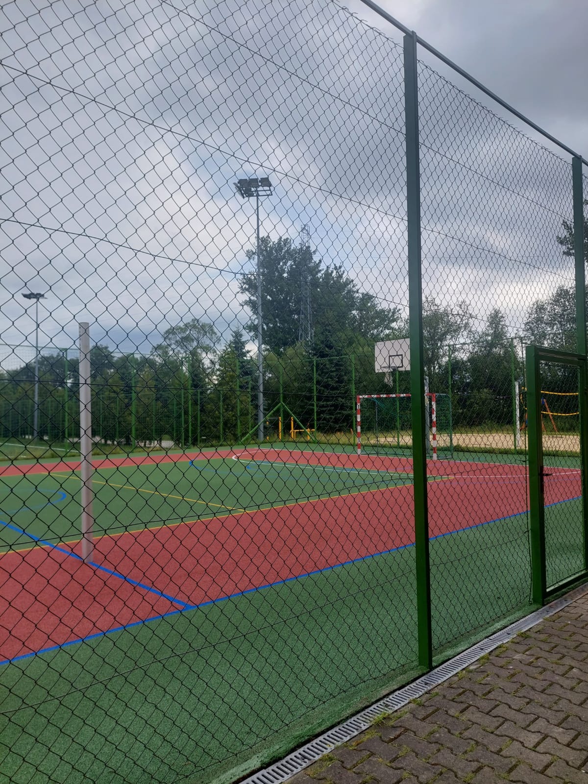 Wielofunkcyjne boisko sportowe z nawierzchnią w kolorach zielonym i czerwonym, ogrodzone siatką z bramką piłkarską i koszem do koszykówki, oświetlone lampami, widok zza ogrodzenia.