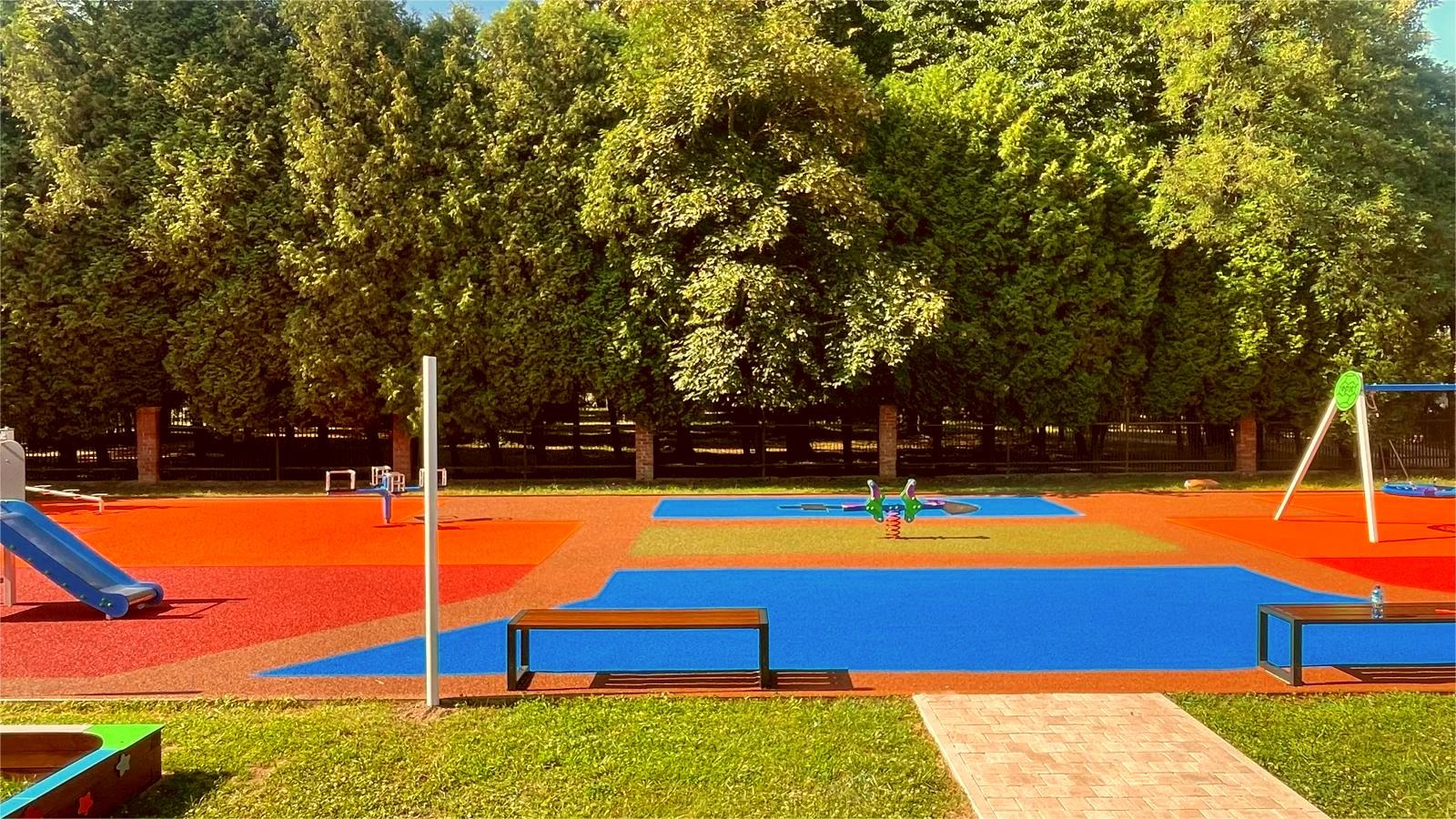 Kolorowy plac zabaw z huśtawkami, zjeżdżalnią i sprężynowcem, otoczony zielenią drzew, z nawierzchnią zróżnicowaną kolorystycznie na strefy.
