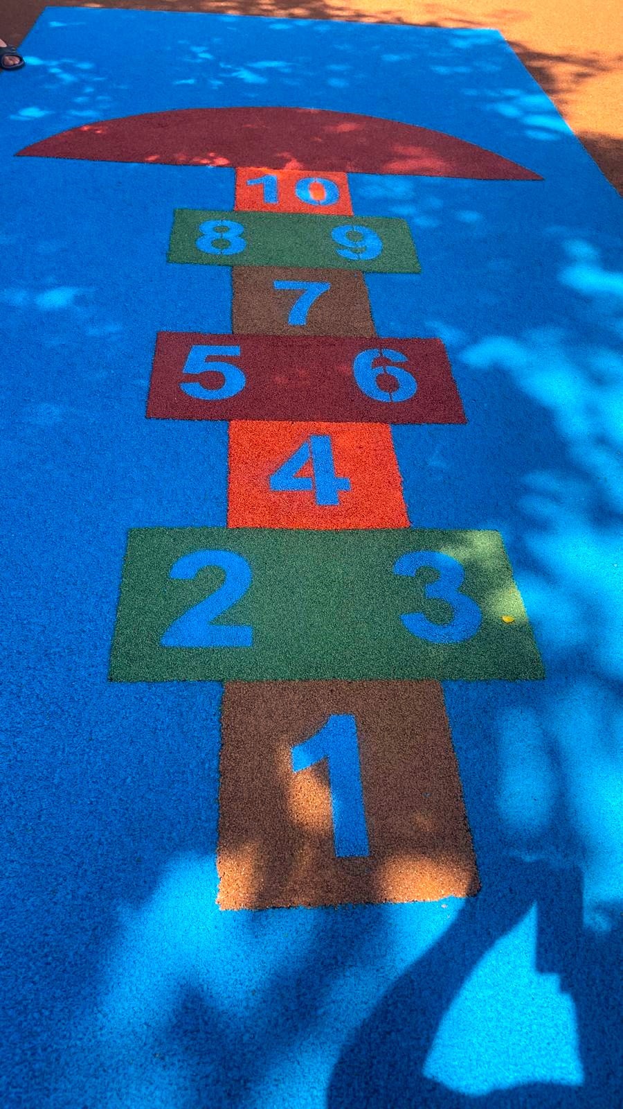 Nawierzchnia placu zabaw z gumowej kostki w kolorze niebieskim, z namalowaną grą w klasy z polami w różnych kolorach i numerami od 1 do 10 w kolorze niebieskim, częściowo zacieniona przez drzewa.