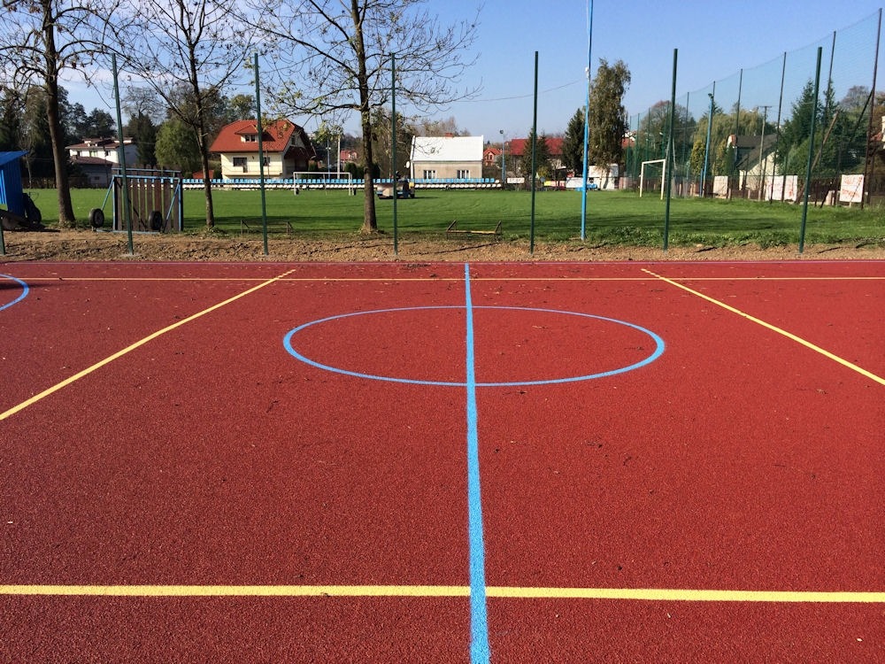 Czerwona nawierzchnia boiska wielofunkcyjnego z niebieskimi i żółtymi liniami, widok na siatkę i bramki piłkarskie w tle.