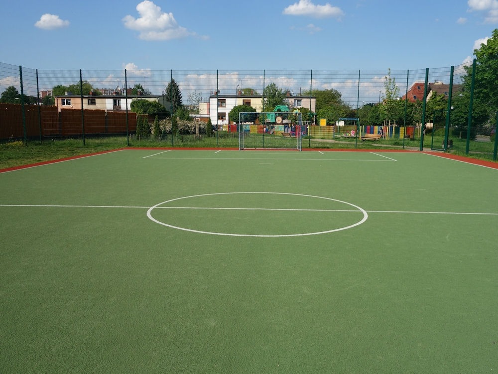 Zielone boisko sportowe z bramkami, otoczone siatką, w tle zabudowa mieszkalna i elementy placu zabaw.