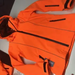 Pomarańczowa kurtka softshell z czarnymi zamkami i elementami odblaskowymi, widoczna metka producenta, leżąca na jasnym tle.