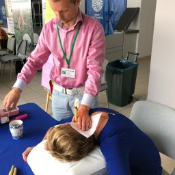 Uzdrowiciel Mariusz Bryk - Mężczyzna w różowej koszuli wykonuje zabieg świecowania uszu u leżącej pacjentki, na stole widoczne akcesoria, w tle banery reklamowe.