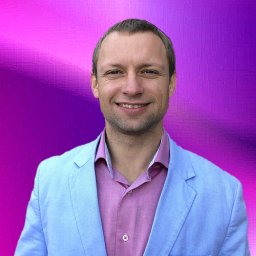 Uzdrowiciel Mariusz Bryk - Uśmiechnięty mężczyzna w błękitnej marynarce i różowej koszuli na rozmytym fioletowo-różowym tle.