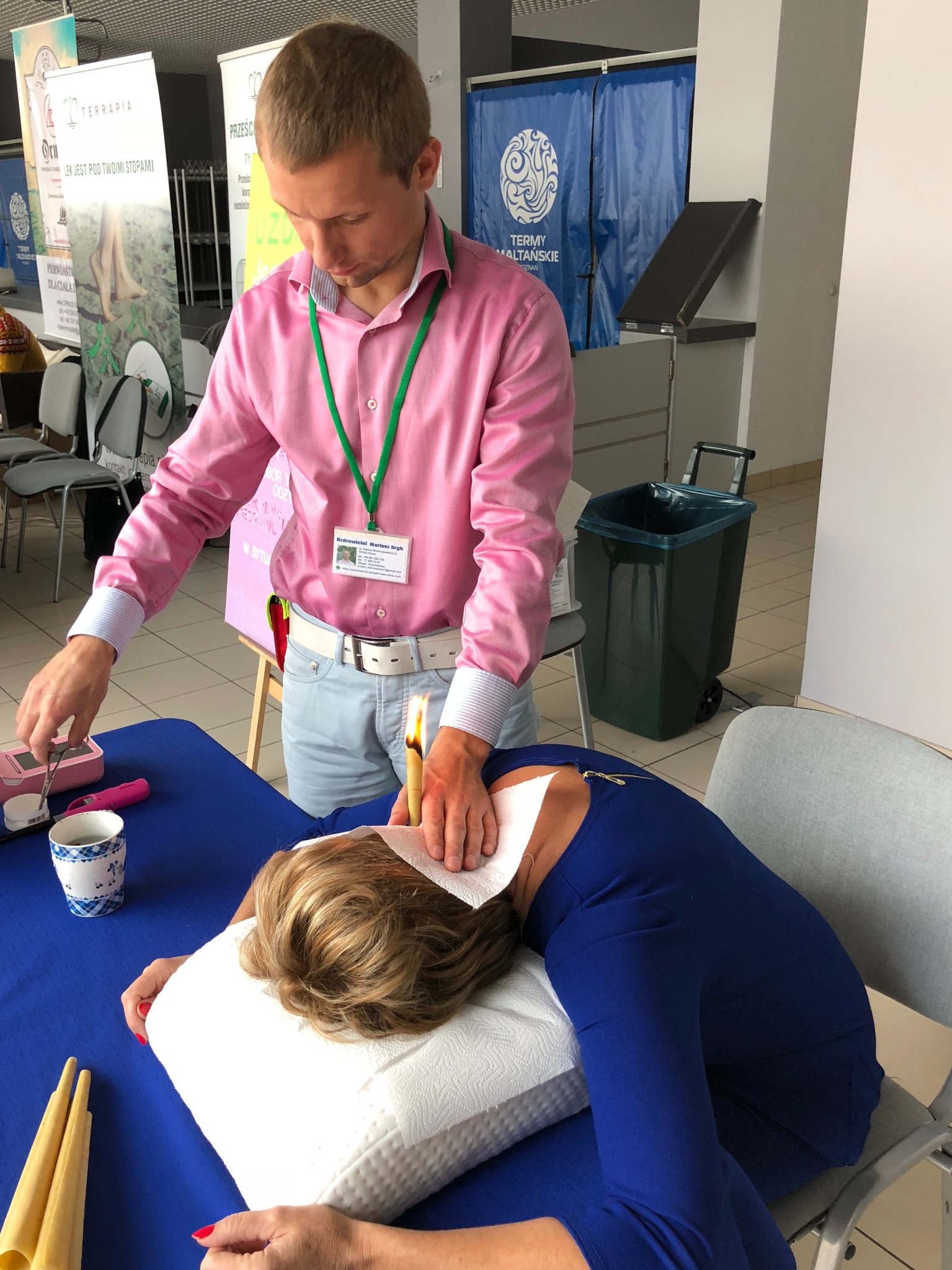 Mężczyzna w różowej koszuli wykonuje zabieg świecowania uszu u leżącej pacjentki, na stole widoczne akcesoria, w tle banery reklamowe.