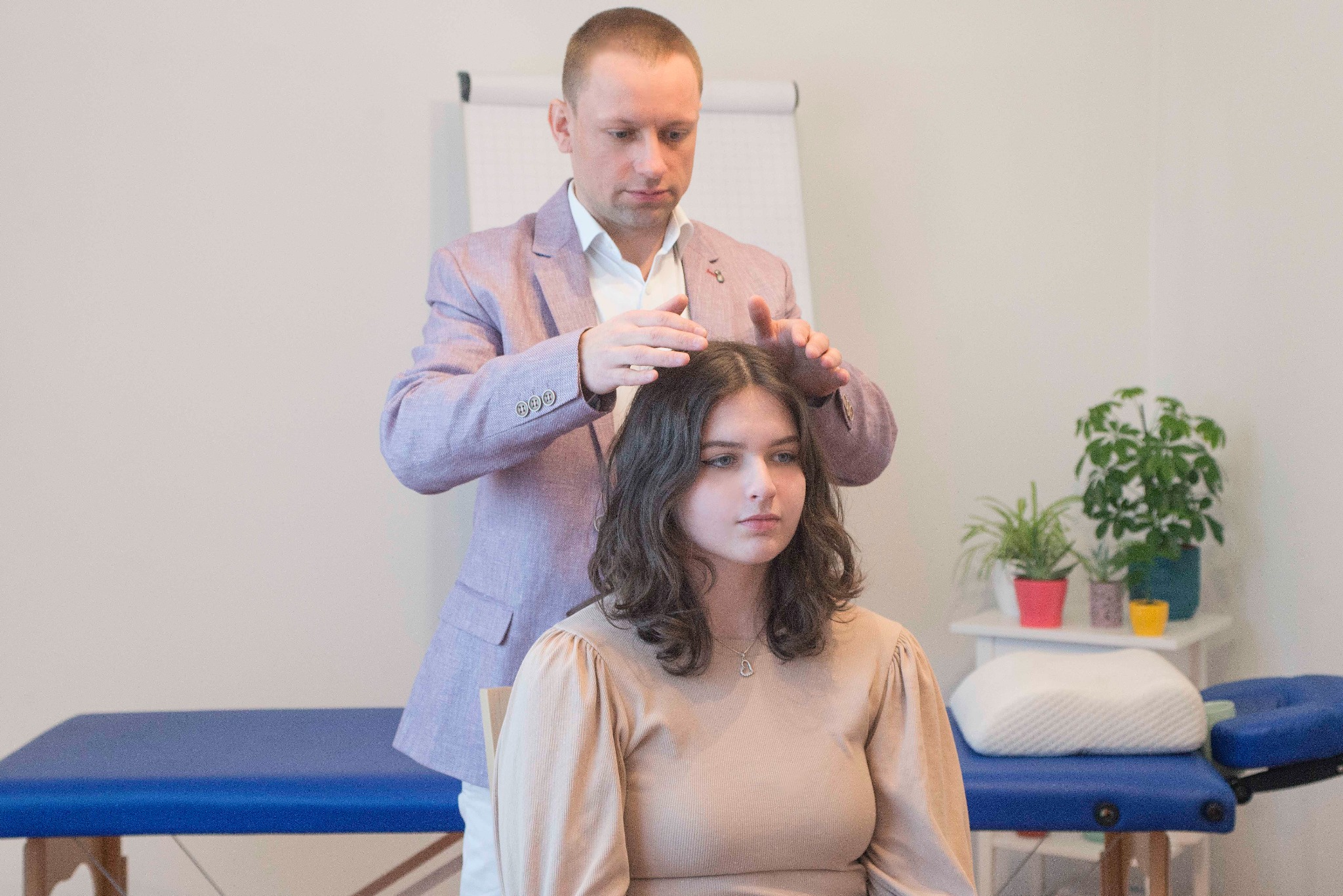 Mężczyzna w jasnofioletowej marynarce delikatnie dotyka głowy siedzącej kobiety podczas sesji terapeutycznej w jasnym pomieszczeniu z roślinami w tle.