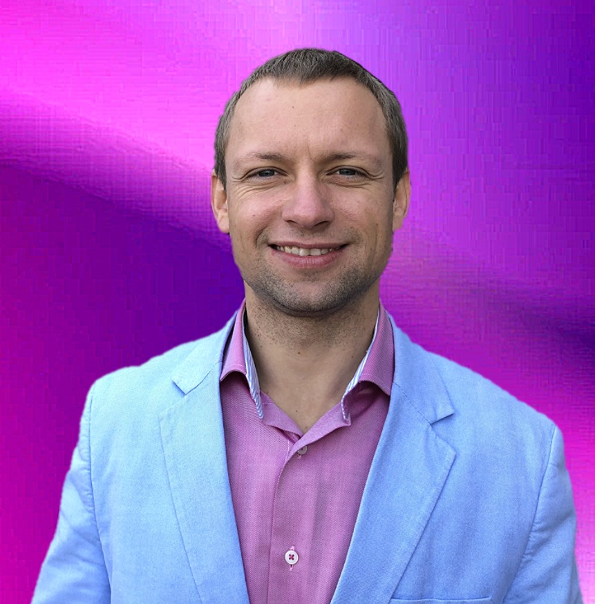 Uśmiechnięty mężczyzna w błękitnej marynarce i różowej koszuli na rozmytym fioletowo-różowym tle.