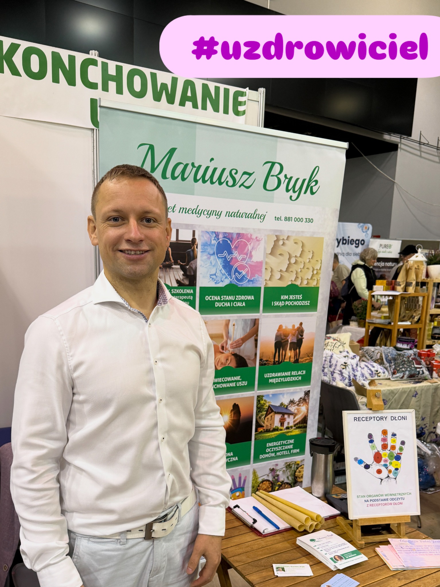 Uśmiechnięty mężczyzna na tle stoiska z napisem 'Mariusz Bryk, gabinet medycyny naturalnej' i hasłem #uzdrowiciel. Widoczne materiały informacyjne i grafika dłoni.