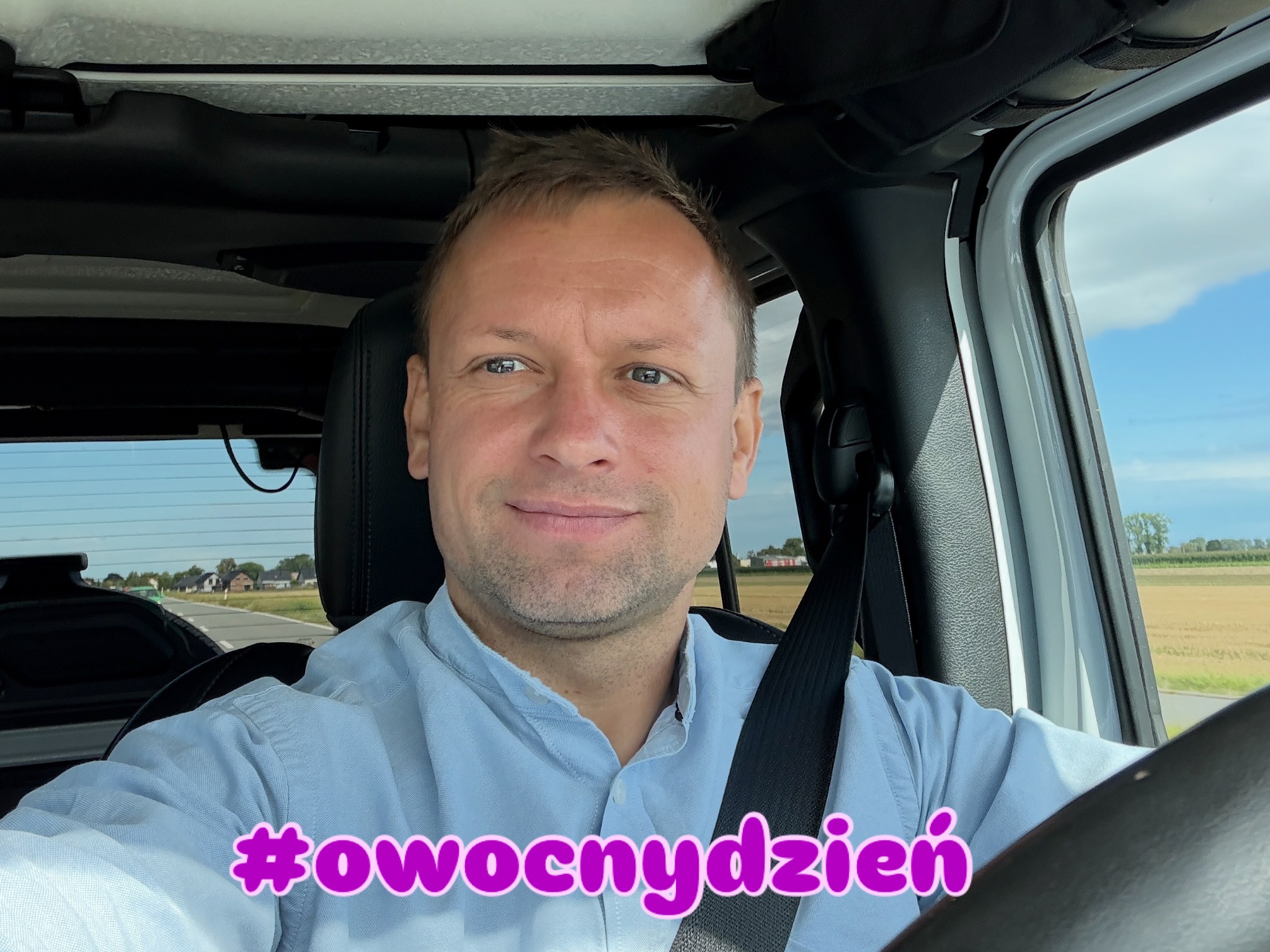 Mężczyzna w błękitnej koszuli, siedzący za kierownicą samochodu, zapięty pasem bezpieczeństwa, z widokiem na krajobraz za oknem i napisem #owocnydzien.