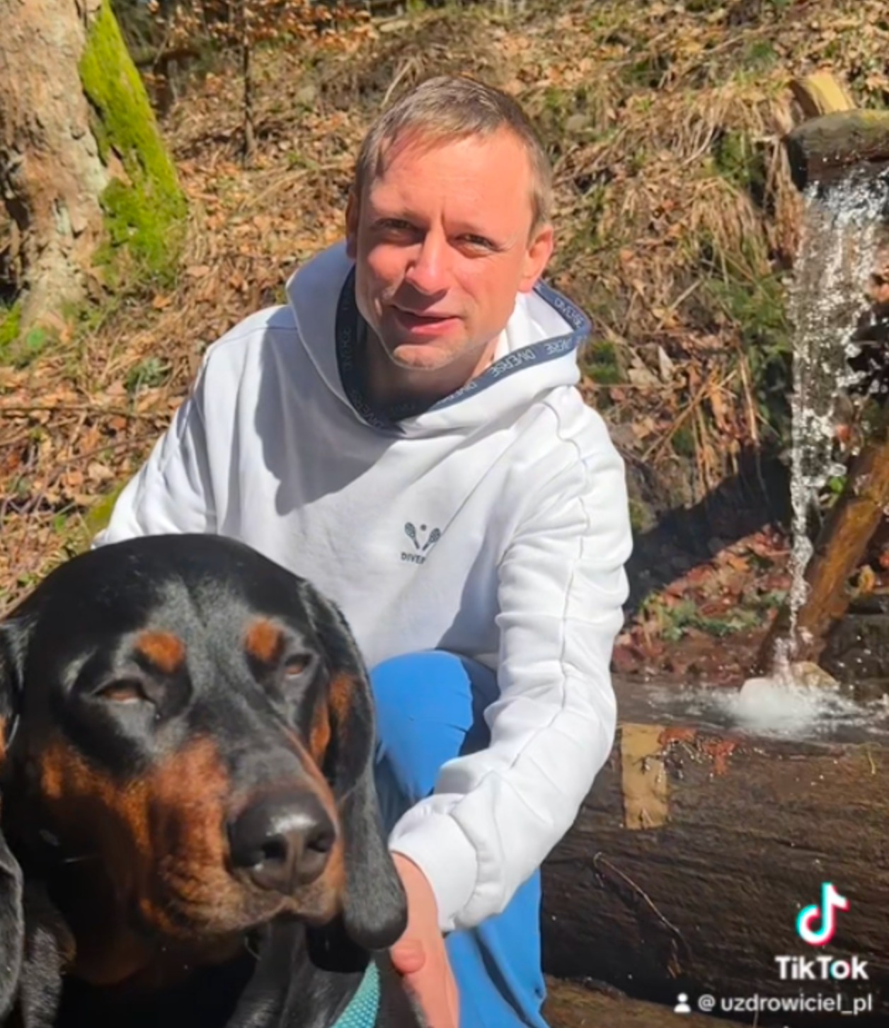 Mężczyzna w białej bluzie z psem rasy gończy polski siedzi na tle leśnego strumienia. Ujęcie z dołu, naturalne światło.