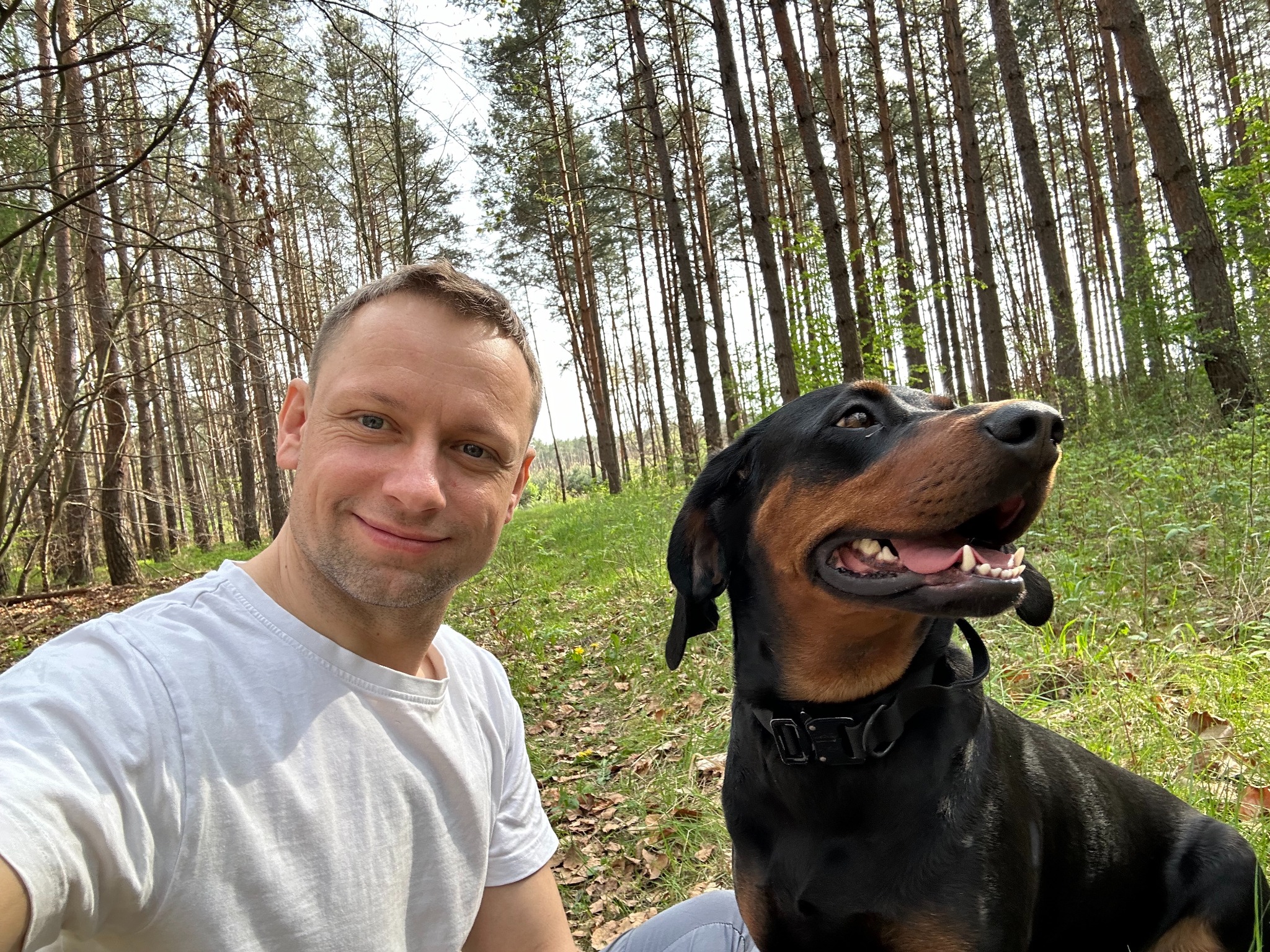 Uśmiechnięty mężczyzna w białej koszulce robi selfie z psem rasy doberman w lesie. W tle wysokie drzewa i zielona trawa.