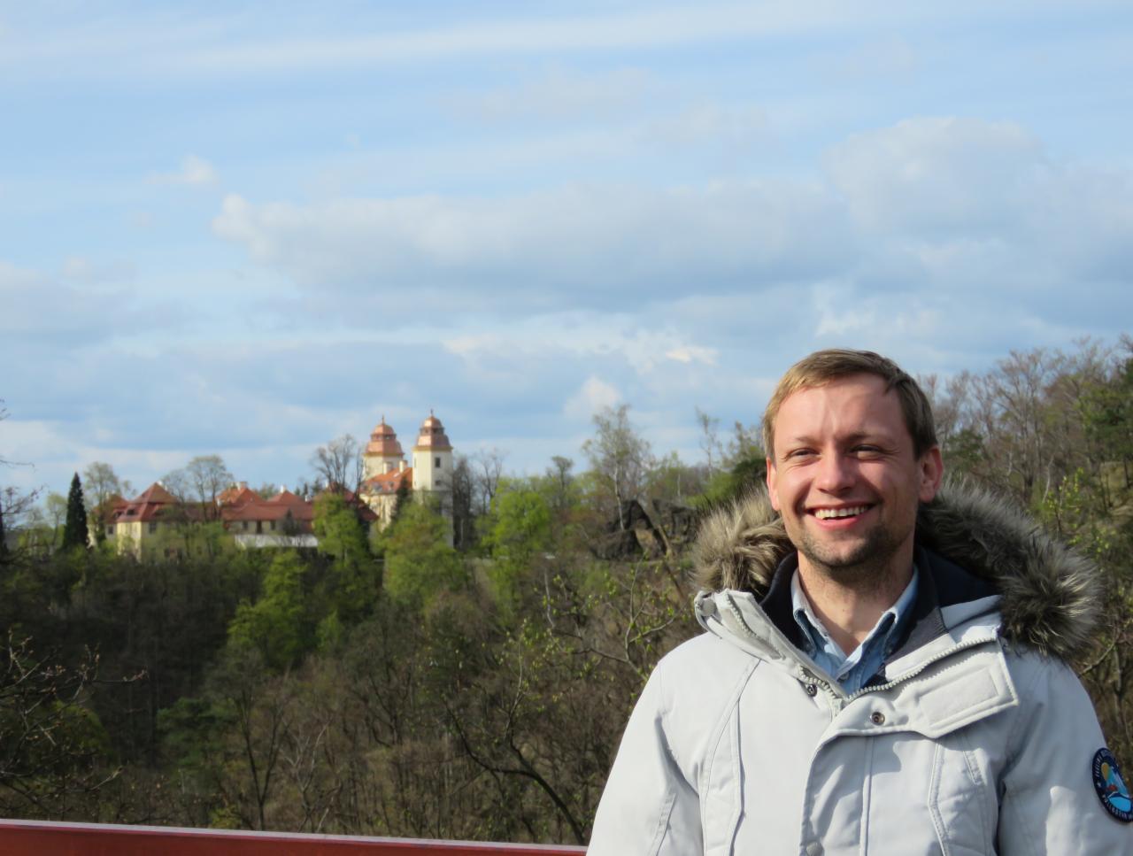 Uśmiechnięty mężczyzna w kurtce z futrzanym kapturem, na tle wzgórza z zamkiem i błękitnego nieba z chmurami.