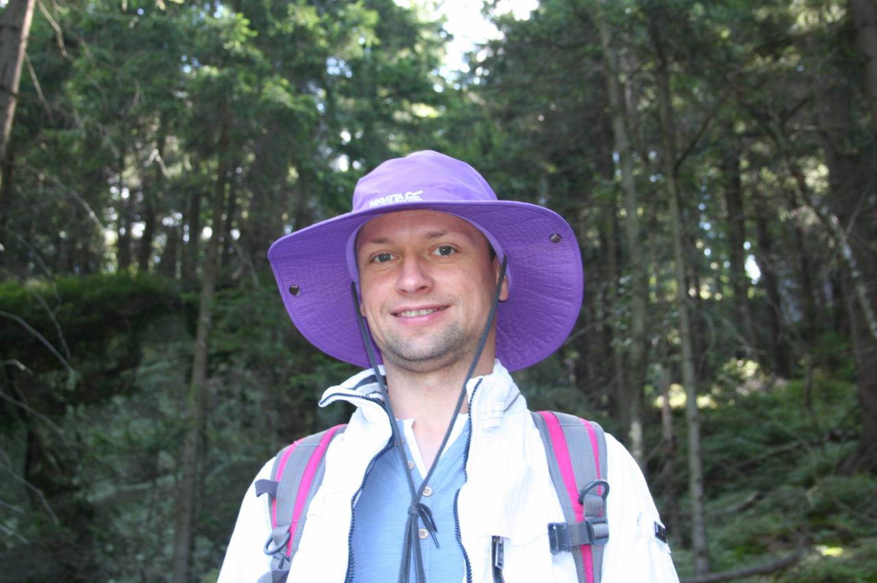 Uśmiechnięty mężczyzna w fioletowym kapeluszu turystycznym i białej kurtce z plecakiem na tle leśnym.