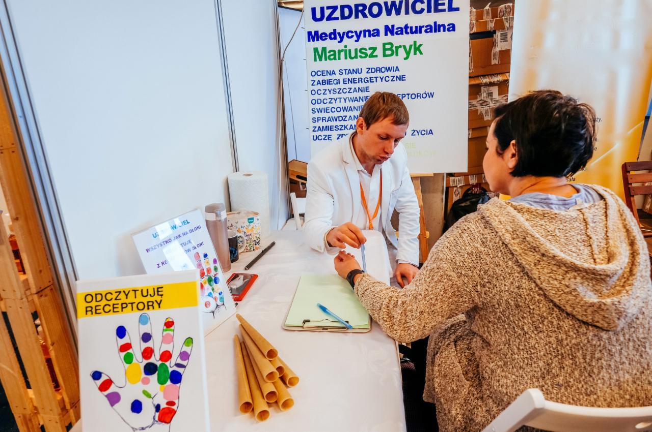 Konsultacja z zakresu medycyny naturalnej: mężczyzna w białym ubraniu sprawdza puls kobiety przy stole, na którym leżą ulotki informacyjne i stożki do świecowania uszu.