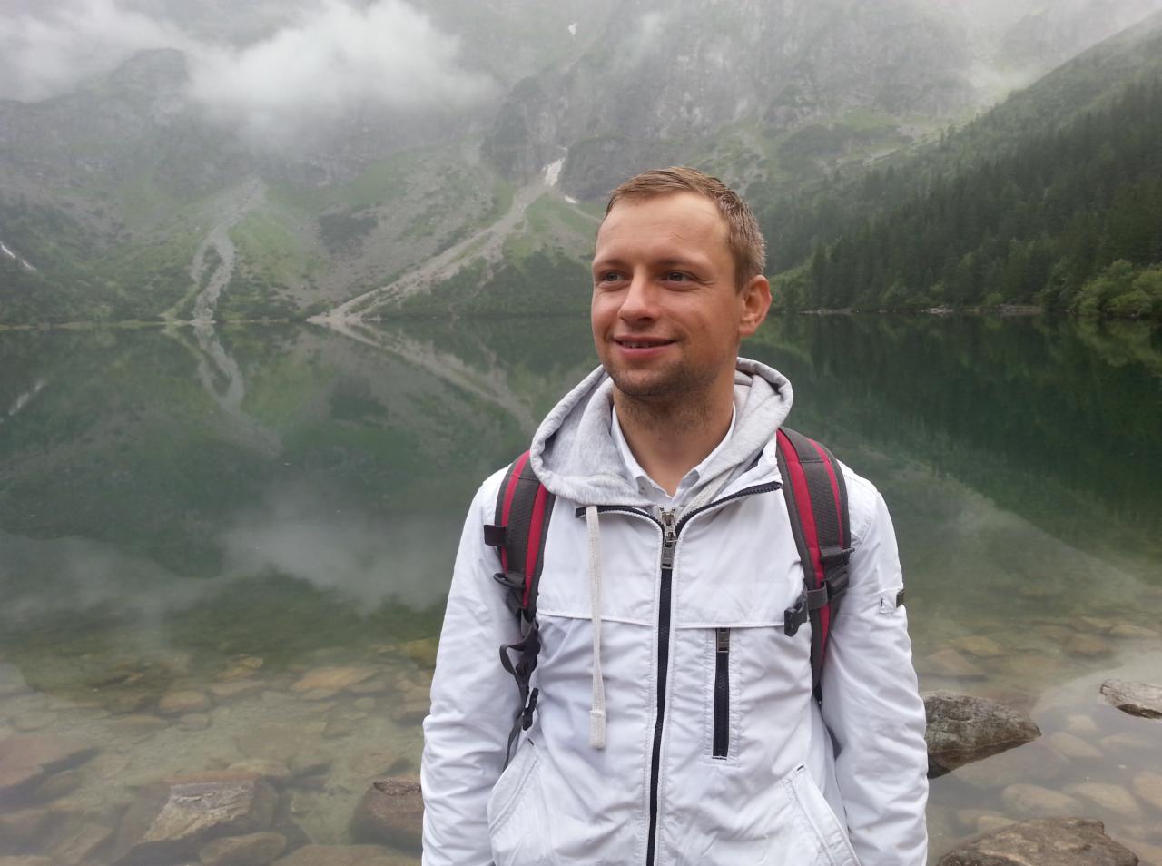 Uśmiechnięty mężczyzna w białej kurtce i z plecakiem na tle górskiego jeziora z mgłą i odbiciem gór w wodzie.