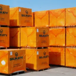 Palety z pomarańczowymi paczkami materiałów budowlanych, owinięte folią, z nadrukowanym logo 'Silikaty Grupa' i danymi kontaktowymi, składowane w magazynie.