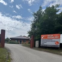 Wjazd do składu budowlanego z widocznym budynkiem biurowym i tablicą informacyjną z ofertą. Ujęcie z dołu, w tle drzewa i błękitne niebo.