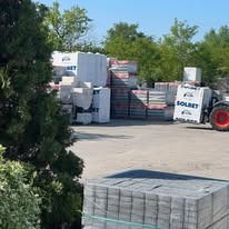 Wózek widłowy przewozi paletę z bloczkami SOLBET na tle stosów materiałów budowlanych na placu składowym w słoneczny dzień. Widoczne drzewa i błękitne niebo.