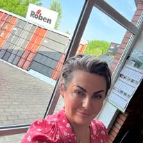 Kobieta robi selfie na tle ekspozycji dachówek Roben, widocznej przez okno. Dachówki w odcieniach czerwieni i brązu, kalendarz na ścianie, jasne tło.