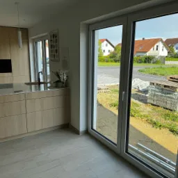 Widok z wnętrza domu przez duże, przesuwne okno balkonowe z białymi ramami. Na zewnątrz widoczny fragment ulicy, domy jednorodzinne i palety z materiałami budowlanymi.