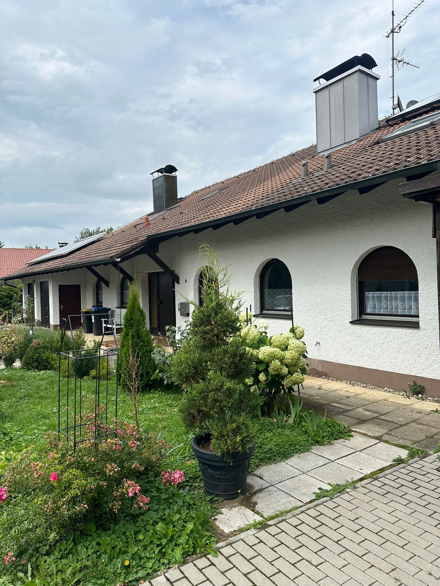 Biały dom z łukowymi oknami, częściowo zasłoniętymi roletami, otoczony zadbanym ogrodem z różami i krzewami hortensji.