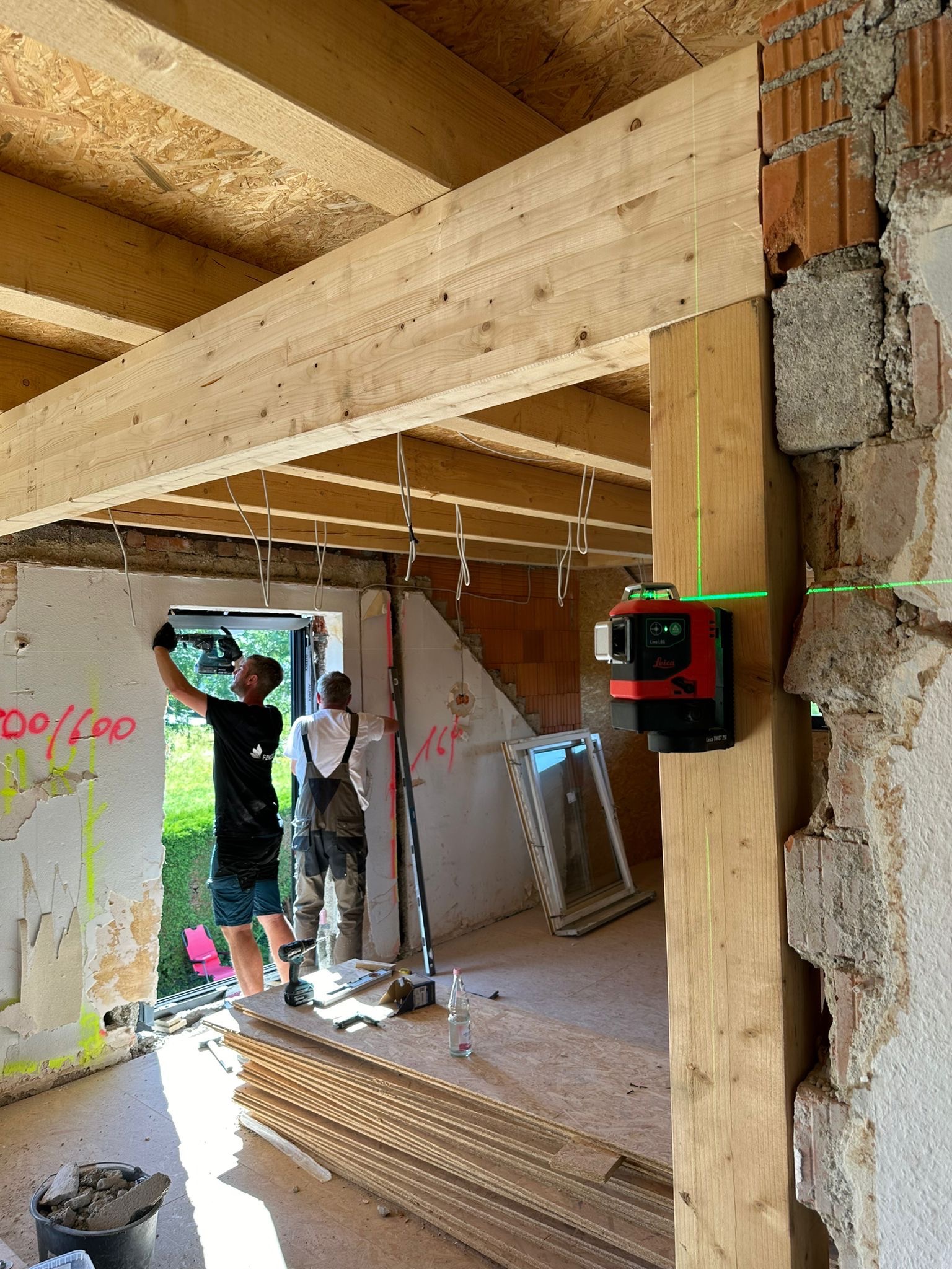 Dwóch monterów instaluje drewniane belki w otworze okiennym w trakcie remontu, używając poziomicy laserowej; widoczne narzędzia i materiały budowlane.