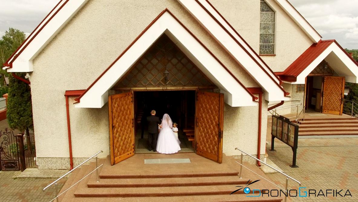 Para młoda wchodzi do kościoła przez otwarte, drewniane drzwi. Ujęcie z drona, widoczny fragment fasady budynku z czerwonymi akcentami.