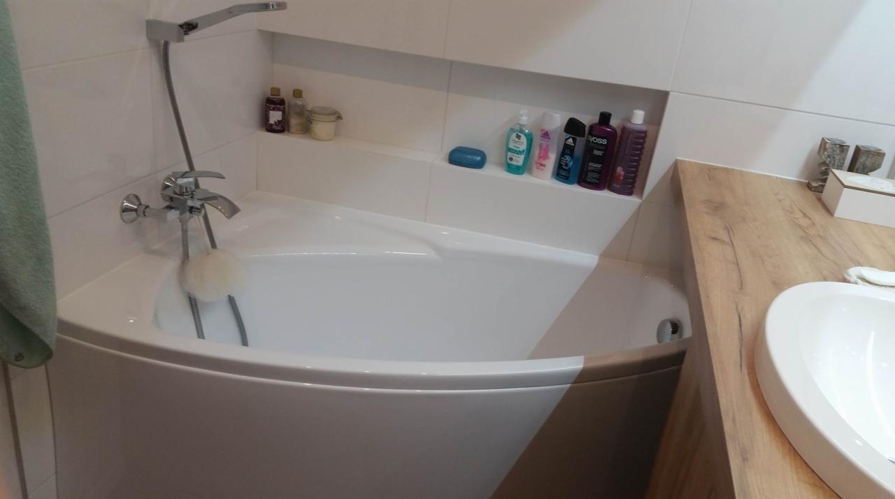 Nowoczesna, narożna wanna z chromowaną armaturą i półką z kosmetykami w jasnej łazience z drewnianym blatem umywalki.