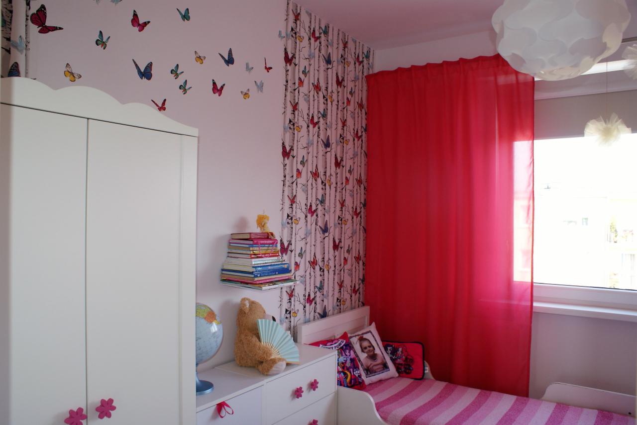 Pokój dziecięcy z białymi meblami, ścianą w motyle, różowymi zasłonami i łóżkiem w różowe paski; na komodzie globus i pluszowy miś z wachlarzem.