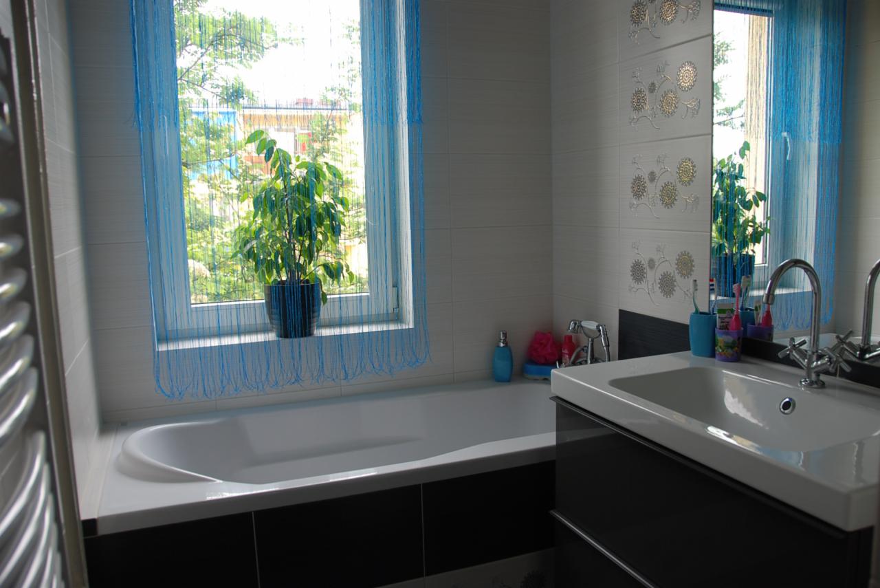 Jasna łazienka z wanną, umywalką w zabudowie i oknem z niebieską firanką sznurkową, na parapecie doniczka z zieloną rośliną, płytki z delikatnym, kwiatowym wzorem
