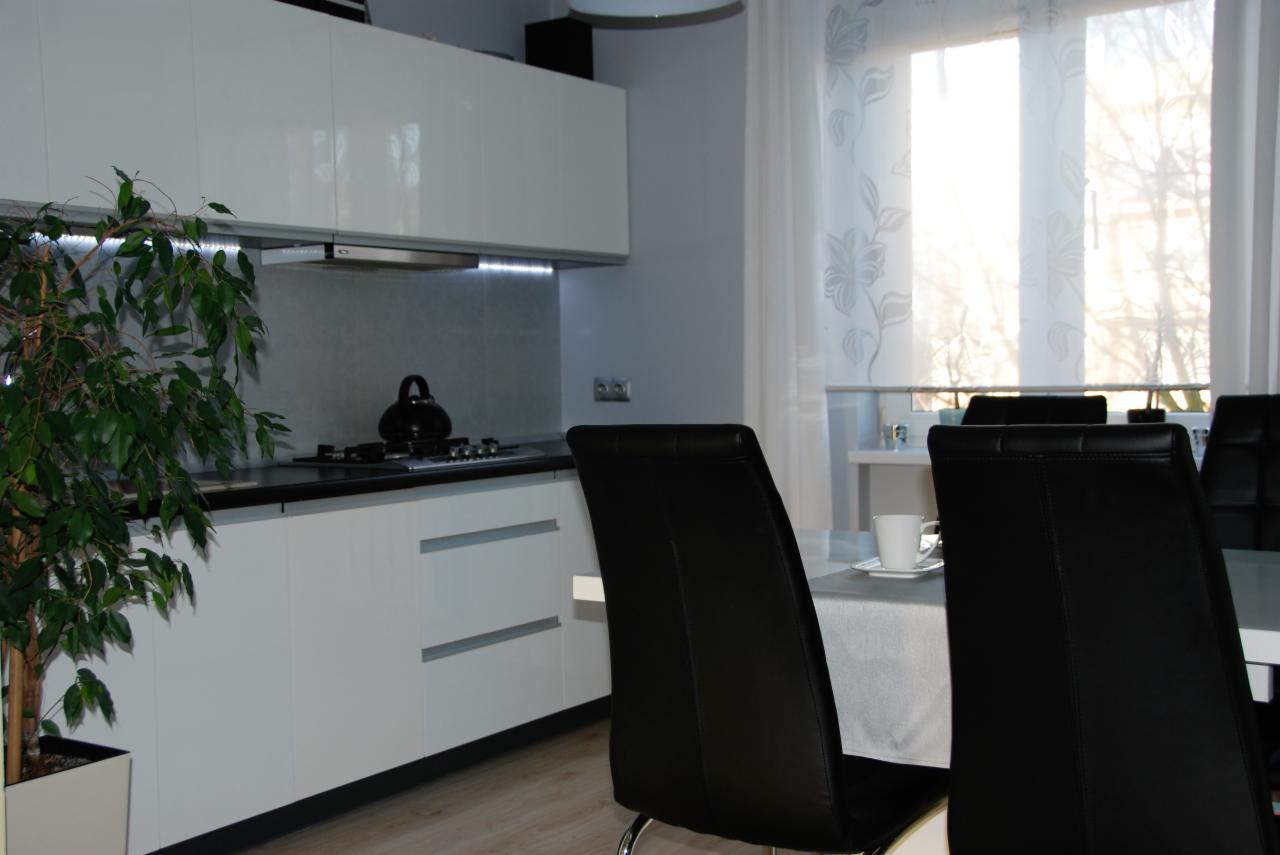 Nowoczesna kuchnia z białymi szafkami, czarnym blatem i oświetleniem LED pod szafkami, widok na stół z czarnymi krzesłami i filiżanką, roślina doniczkowa w lewym rogu.