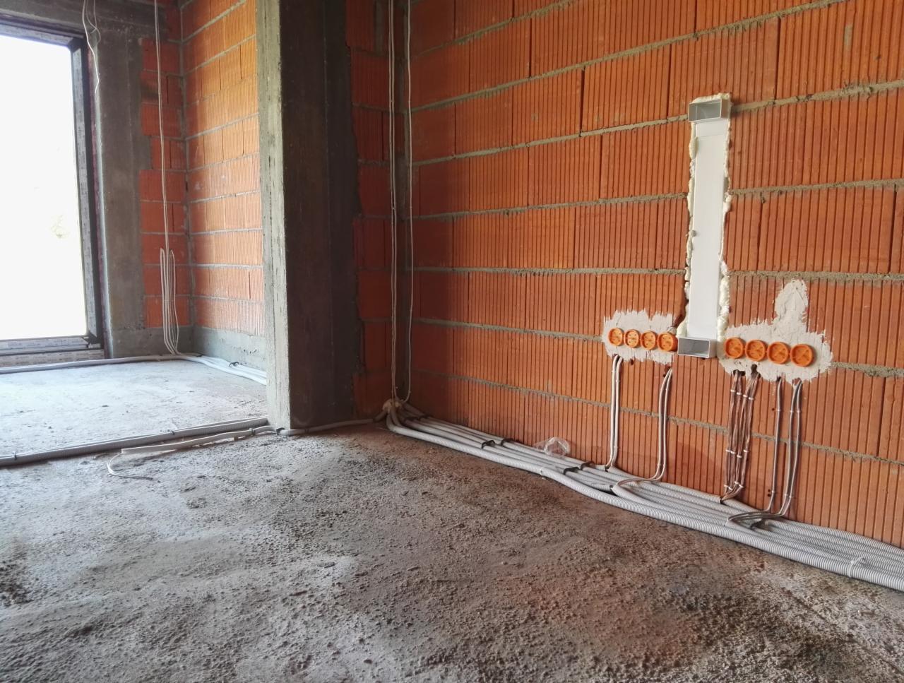 Wnętrze budynku w stanie surowym otwartym z widoczną instalacją elektryczną w ścianie z cegły ceramicznej i przewodami w podłodze.
