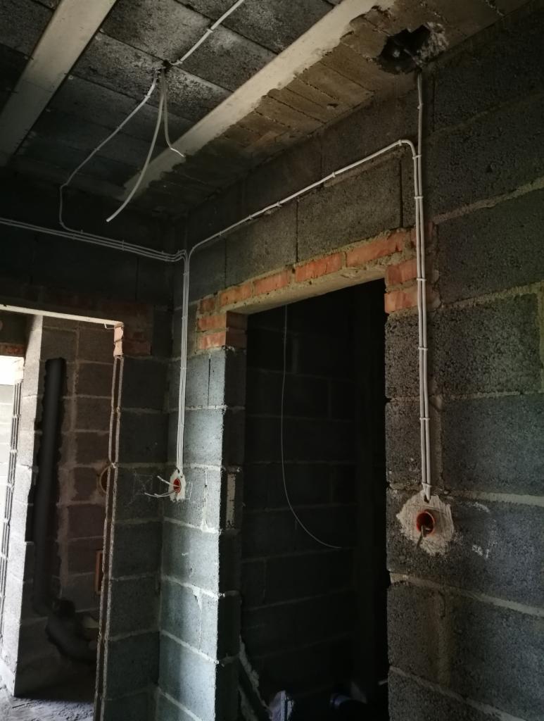 Surowy stan budynku w trakcie prac instalacyjnych: widoczne szare bloczki, ceglane obramowania drzwi, oraz poprowadzone białe kable elektryczne w peszlach i puszki instalacyjne.