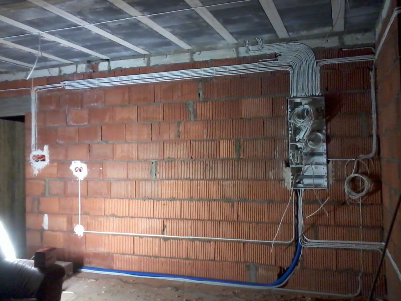 Instalacja elektryczna w budynku w trakcie budowy: widoczne kable w białych rurkach prowadzone po ścianie z czerwonej cegły do skrzynki rozdzielczej, sufit w stanie surowym.