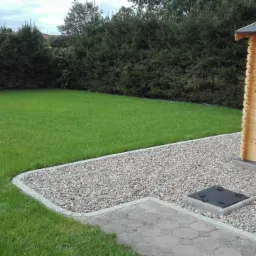 Zadbany trawnik z równo przystrzyżoną trawą, drewniany domek ogrodowy z okiennicą, ścieżka z kostki brukowej i kamienny obrzeże. W tle wysokie, gęste tuje.