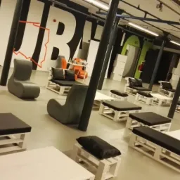 Przestrzeń eventowa z meblami z palet: białe stoliki i siedziska z czarnymi poduchami, szare fotele, na ścianie czarno-biały mural z elementami czerwieni, oświetlenie przemysłowe.