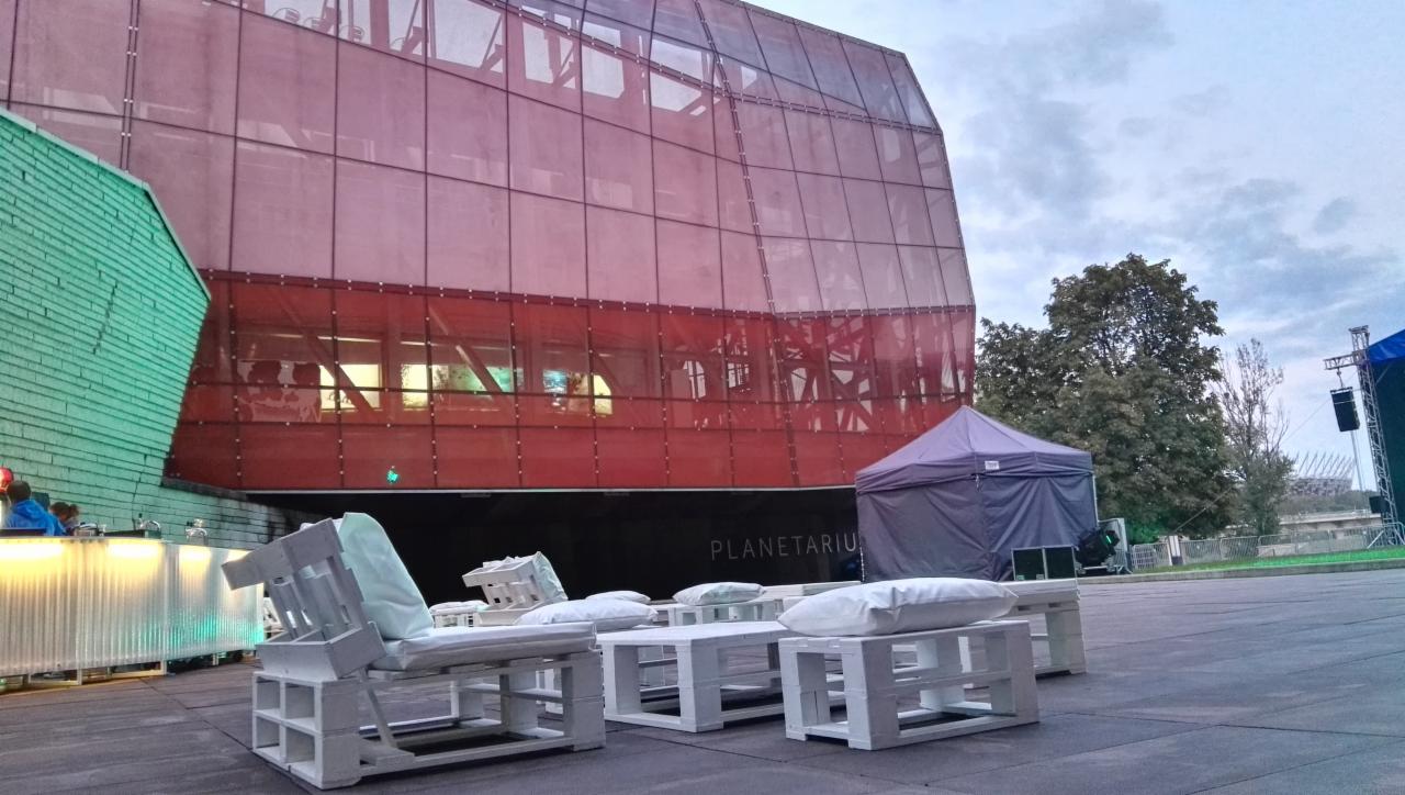 Przestrzeń eventowa na zewnątrz budynku Planetarium z meblami z palet, oświetlonym barem i namiotem w tle, widoczny Stadion Narodowy.