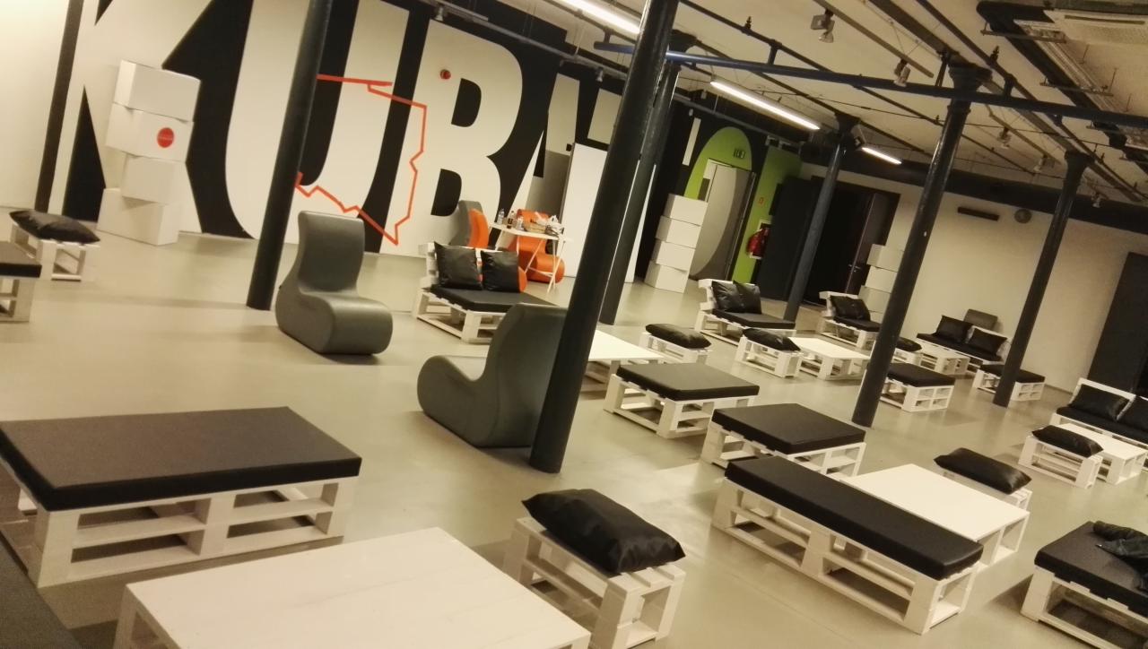 Przestrzeń eventowa z meblami z palet: białe stoliki i siedziska z czarnymi poduchami, szare fotele, na ścianie czarno-biały mural z elementami czerwieni, oświetlenie przemysłowe.