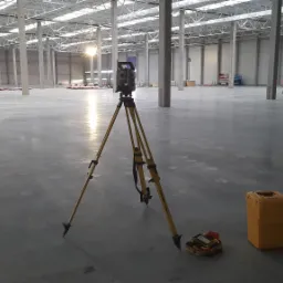 Geodezyjny tachimetr elektroniczny Trimble na statywie w surowym, betonowym wnętrzu hali magazynowej z widocznymi słupami nośnymi i osobami w oddali, obok żółta skrzynka transportowa.