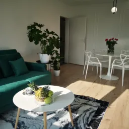 Jasne wnętrze z zieloną sofą, okrągłym stolikiem z owocami, dywanem w marmurkowy wzór, okrągłym stołem z kwiatami i białymi krzesłami w stylu skandynawskim, oświetlone naturalnym światłem