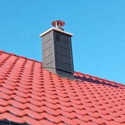 TECTUM PiotrGrzewinski - Czerwony dach z ceramicznej dachówki z czarnym kominem wentylacyjnym zwieńczonym wiatraczkiem na tle błękitnego nieba w słoneczny dzień.