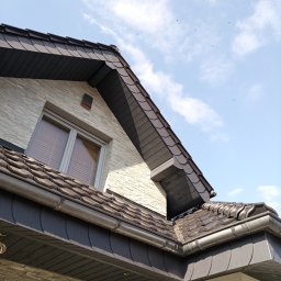 TECTUM PiotrGrzewinski - Nowoczesny dach z ciemnej dachówki ceramicznej i szarymi rynnami, widok z dołu na tle błękitnego nieba z chmurami. Elewacja budynku wykończona kamieniem.